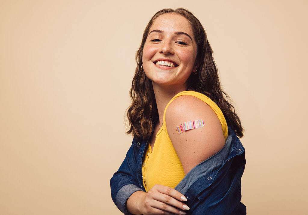 Portrait of a female smiling after getting a vaccine. Woman holding down her shirt sleeve and showing her arm with bandage after receiving vaccination.